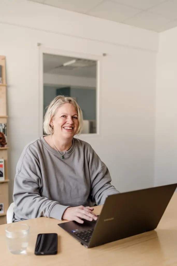 Antje Schwirz am Schreibtisch im Büro vor dem Laptop, lachend in die Kamera blickend