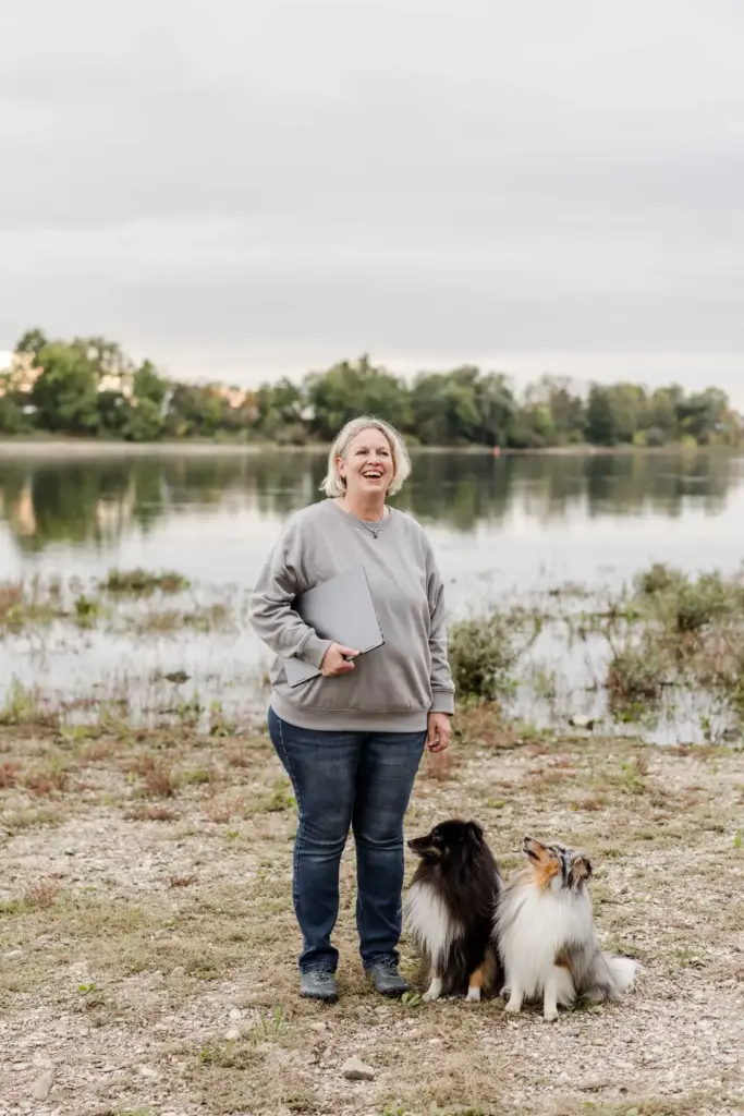 Antje Schwirz an der Donau, lachend, einen Laptop unter der Hand, zwei Hunde zu ihren Füßen